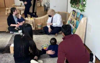 Brightly lit child reading corner at Monkey Puzzle Day Nurseries with books, soft rugs, and a group of parents and children engaged in storytelling session, creating a warm, educational environment for early childhood development.