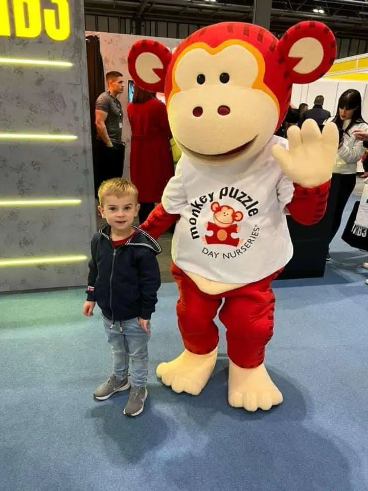 Monkey Puzzle Day Nurseries mascot in a colorful monkey costume at an event with children and parents.
