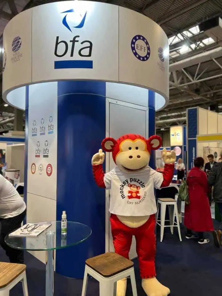 Monkey Puzzle Day Nurseries promotional mascot at a trade show booth, dressed as a friendly monkey in a white tee, encouraging early childhood education and daycare services.