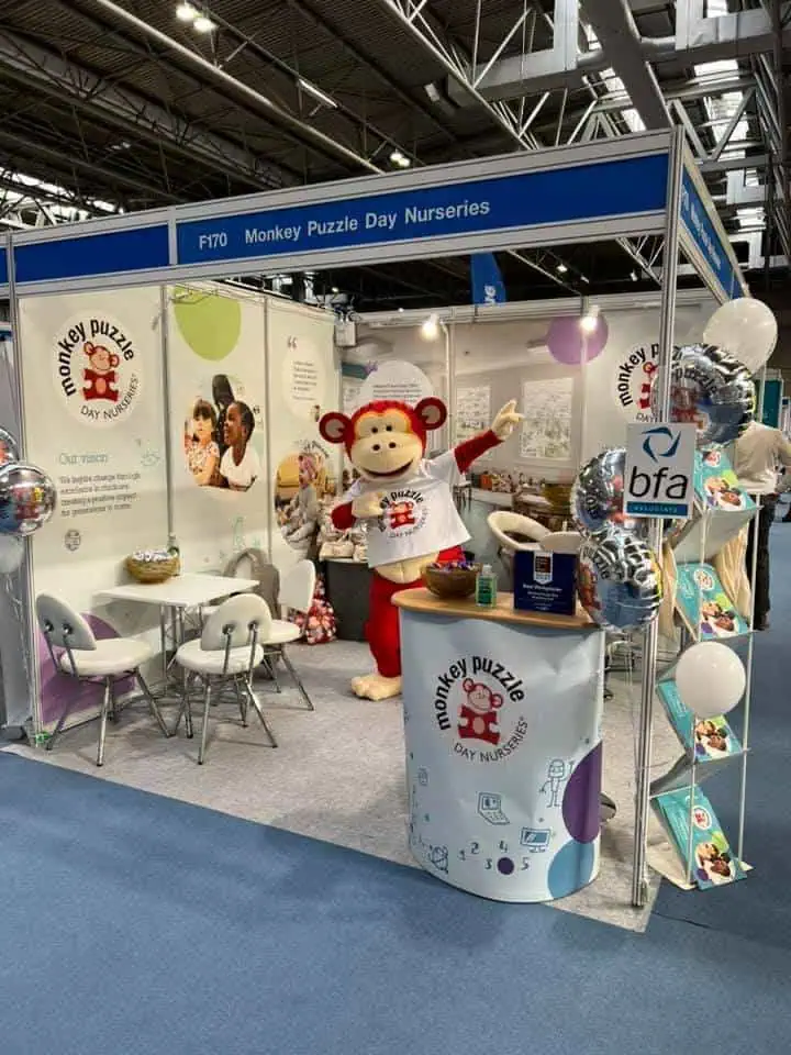 Monkey puzzle day nurseries booth at exhibition, featuring a mascot in a monkey costume, promotional materials, and seating area, highlighting early childhood education and daycare services for young children.