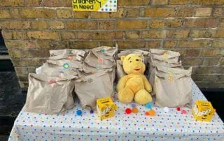 Brown paper goody bags with colorful stickers, a yellow Winnie the Pooh plush toy, and small pom-poms on a star-patterned tablecloth, supporting children in need charity event at Monkey Puzzle Day Nurseries.