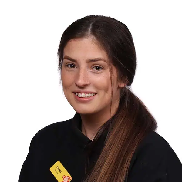 Young female nursery staff member at Monkey Puzzle Day Nurseries smiling, wearing a black uniform with a yellow name badge, in a professional and friendly childcare environment.