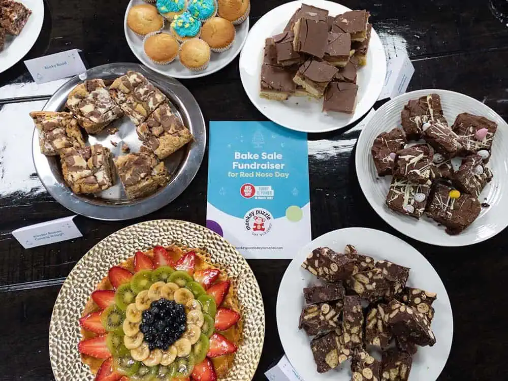 Cakes ready for Comic Relief sale Vegan chocolate brownies on a white plate with a variety of other baked goods and a fruit platter, displayed on a wooden table at a Monkey Puzzle Day Nurseries bake sale fundraiser.