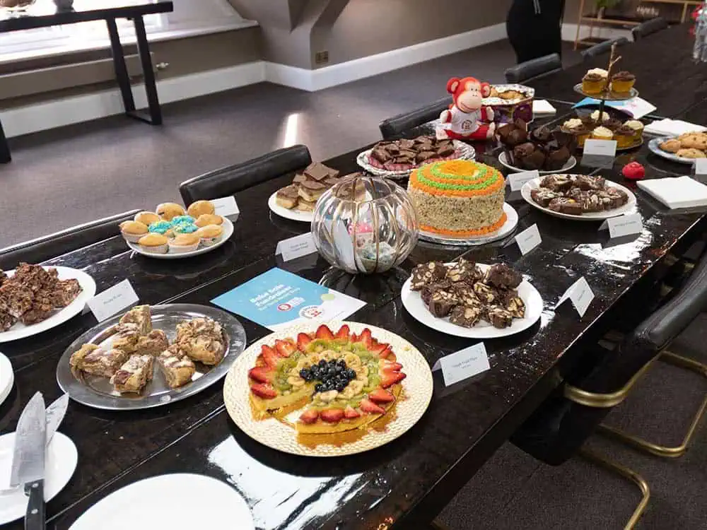 Cakes ready for Red Nose Day sale A variety of beautifully decorated cakes and sweet treats displayed on a dark conference table for a celebration event at Monkey Puzzle Day Nurseries.