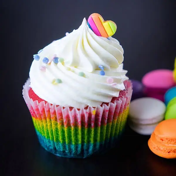 Rainbow cupcake with colorful sprinkles and a heart-shaped topping, surrounded by other macarons, on a dark background. Perfect for children's celebrations and catering highlights at Monkey Puzzle Day Nurseries.