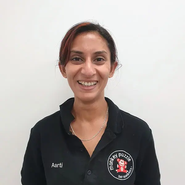 Friendly female nursery practitioner smiling, wearing a Monkey Puzzle Day Nurseries uniform with embroidered logo and name tag, promoting quality childcare services.