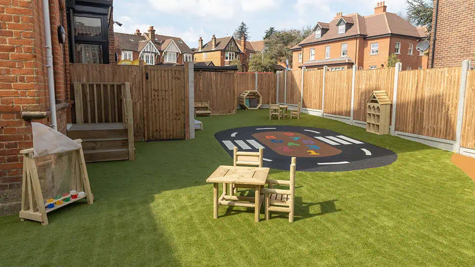 Colorful outdoor play area at Monkey Puzzle Day Nurseries, featuring a soft artificial grass surface, children's wooden tables and chairs, and engaging playground markings for early childhood fun and development.