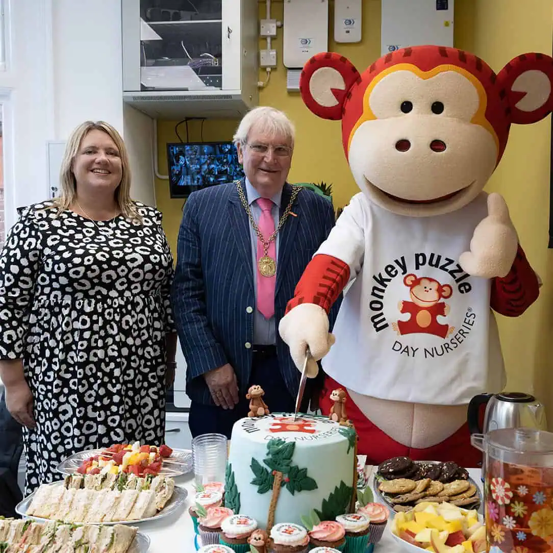 Mayor of Guildford Dennis Booth and Monkey Puzzle nursery manager Cheerful celebration at Monkey Puzzle Day Nurseries with staff, the mascot, and a decorated cake featuring the nursery's logo during a special event.