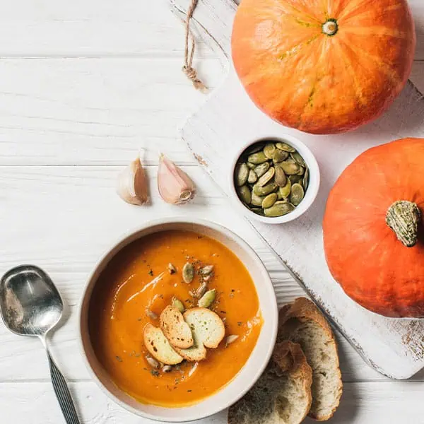 Creamy pumpkin soup garnished with croutons and pumpkin seeds, served with fresh bread, garlic, and pumpkins on a rustic white wooden table at Monkey Puzzle Day Nurseries.