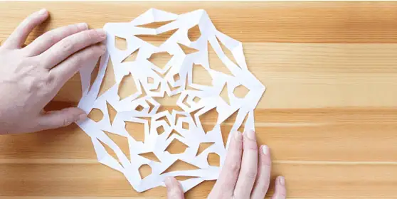 Paper snow flakes – Step 8 Intricate paper snowflake craft being made by hands on a wooden surface, showcasing creative activities at Monkey Puzzle Day Nurseries.