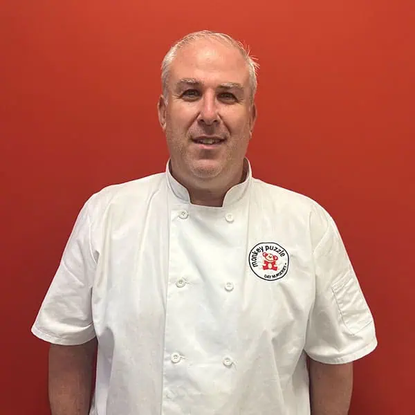 Chef in white uniform with Monkey Puzzle Day Nurseries logo on chest, standing against a bright orange background, promoting childcare and early education services.