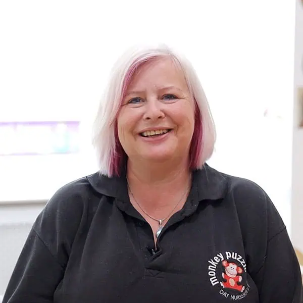 Friendly female daycare staff member at Monkey Puzzle Day Nurseries wearing a black polo shirt, smiling, in a child care environment.