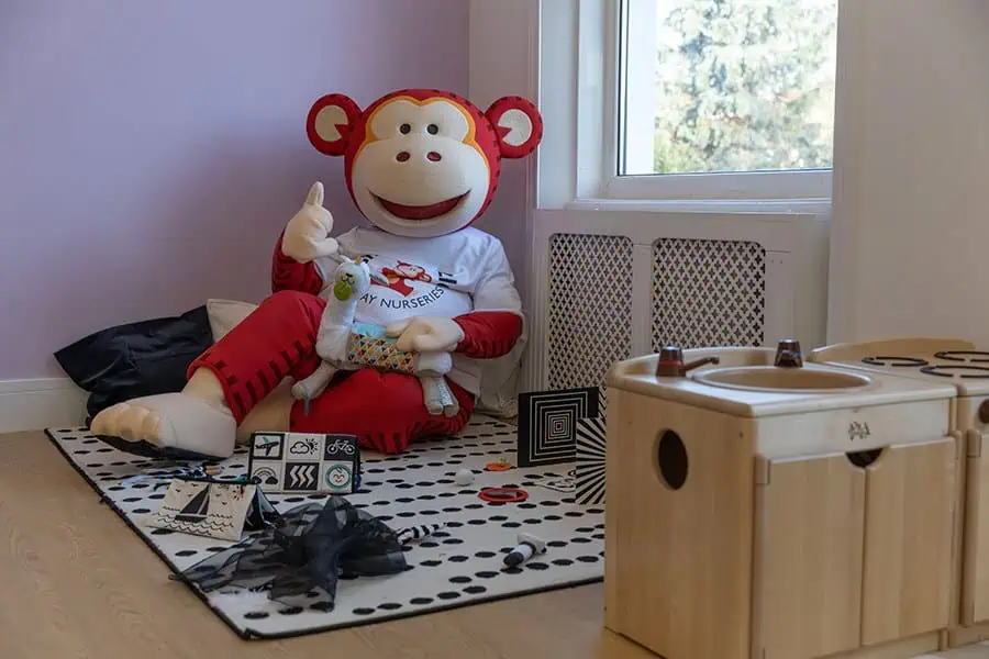 Marvin the Monkey at the opening of Monkey Puzzle Hither Green Colorful monkey mascot sitting on a black and white polka dot rug, holding a plush duck toy at Monkey Puzzle Day Nurseries, creating a fun and welcoming environment for young children.