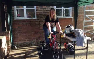 Staff taking turns on the bike ride Bike riding activity at Monkey Puzzle Day Nurseries, outdoor play area with a smiling woman on a stationary bike under a canopy. Engaging physical activity for children, promoting health and fun.