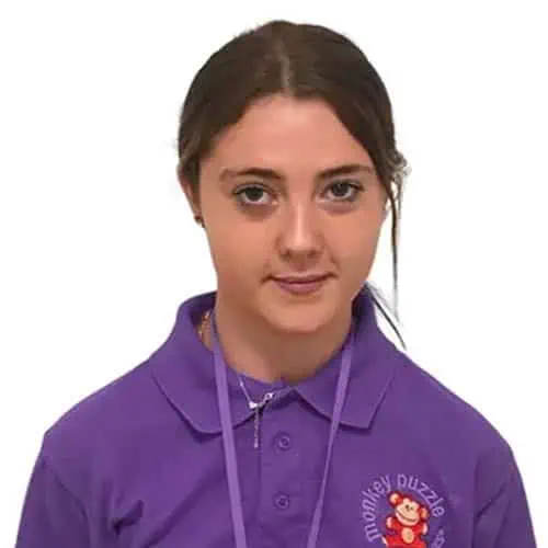 Friendly young woman in purple uniform with Monkey Puzzle logo, smiling confidently, representing a caring childcare professional at Monkey Puzzle Day Nurseries.