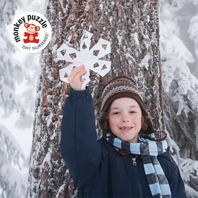 Cute young boy holding a snowflake craft in winter outdoor setting at Monkey Puzzle Day Nurseries, emphasizing early childhood education and outdoor activities.