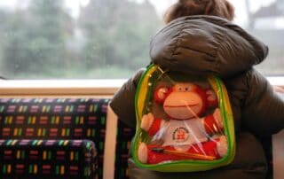 Backpack featuring a Monkey Puzzle Day Nursery plush toy on a child's back while traveling on a train.