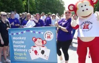 Colorful outdoor parade scene featuring staff and mascot from Monkey Puzzle Day Nurseries, promoting early childhood education and community engagement.