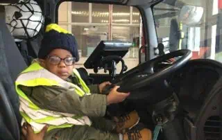 Child sitting in a truck driver's seat wearing a safety vest and beanie, showcasing activities at Monkey Puzzle Day Nurseries for early childhood development and fun learning experiences.