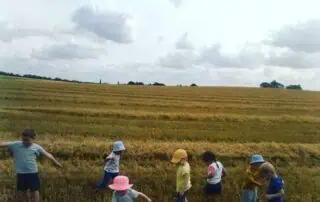 Kids exploring a field near a farmland during daytime, wearing colorful hats and casual clothes, engaging in outdoor activity at Monkey Puzzle Day Nurseries.