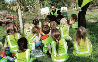 Children's outdoor learning activity at Monkey Puzzle Day Nurseries, sitting on grass in high-visibility vests while listening to a teacher's storytelling session in a garden setting.