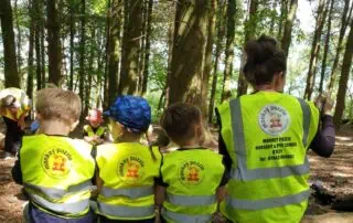 Children and preschool staff at Monkey Puzzle Day Nurseries enjoying outdoor forest learning activity wearing bright yellow vests. The session promotes early education, nature exploration, and outdoor play for preschoolers.