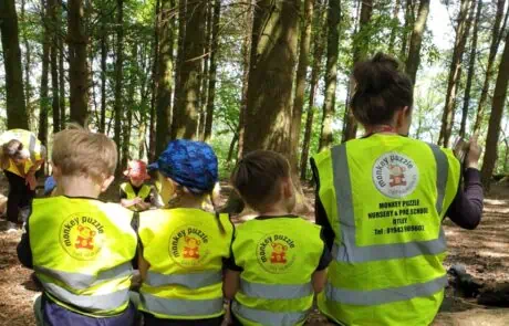 Visits to the woods and local nature spots Children and preschool staff at Monkey Puzzle Day Nurseries enjoying outdoor forest learning activity wearing bright yellow vests. The session promotes early education, nature exploration, and outdoor play for preschoolers.