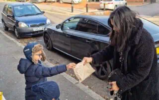 Friendly woman handing a paper bag to a young boy with glasses and a backpack, during an outdoor event on a city street, with parked cars and buildings in the background, promoting community engagement at Monkey Puzzle Day Nurseries.
