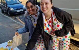 Bright smiling nursery staff wearing colorful polka dot outfits handing out treats outdoors, promoting playful and engaging childcare at Monkey Puzzle Day Nurseries in a lively neighborhood setting.