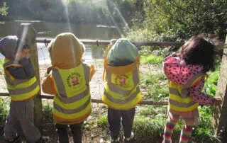 Children exploring nature at Monkey Puzzle Day Nurseries, wearing high-visibility vests near the river, engaging in outdoor learning and environmental activities for preschool development.
