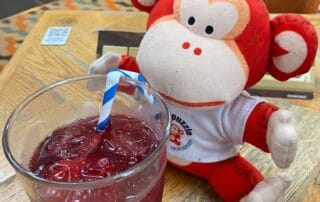 Colorful monkey plush toy sitting at a wooden table with a glass of red beverage with ice and a striped straw in a cozy nursery or cafe setting.