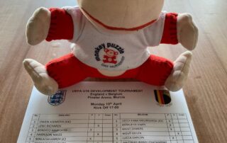 Monkey Puzzle Day Nurseries plush monkey toy wearing a white T-shirt, sitting on a table near a printed football match schedule, representing a fun and engaging environment for early childhood education.