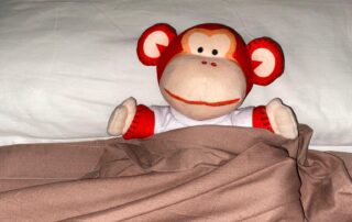 Cute monkey plush toy dressed in a white shirt, sitting on a bed with beige bedding at Monkey Puzzle Day Nurseries.