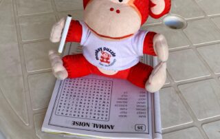 Monkey Puzzle Day Nurseries plush monkey toy sitting on a table with a children's workbook and a pen, promoting engaging activities for early childhood development. Perfect for childcare and preschool learning environments.