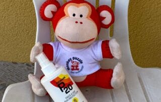 Bright orange monkey plush toy from Monkey Puzzle Day Nurseries holding sunscreen, sitting on a white outdoor chair against a yellow wall, promoting child safety and sun protection.