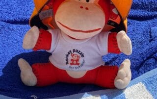 Soft plush monkey toy wearing a Monkey Puzzle Day Nurseries t-shirt and an orange helmet, sitting on a blue towel, symbolizing early childhood play and nursery care.