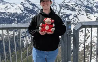 Red monkey plush toy held by smiling woman on mountain viewpoint, snowy peaks in background, outdoor adventure, mountain scenery, child's toy, travel, nature, outdoor activity, family trip, scenic landscape.