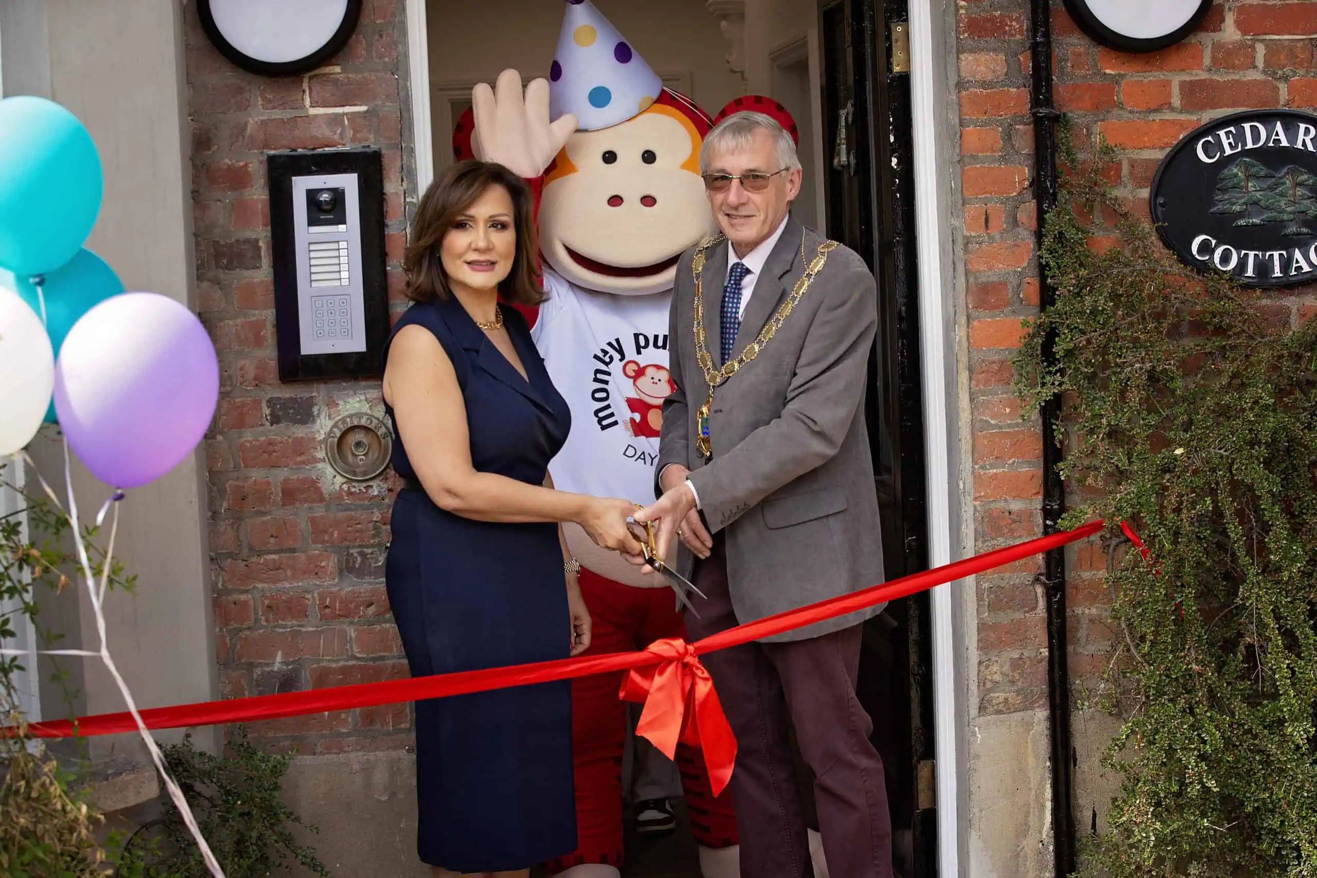 MonkeyPuzzle_Epsom_189 Colorful grand opening of Monkey Puzzle Day Nurseries with ribbon cutting ceremony, featuring staff, mayor, and mascot outside nursery entrance with balloons and decorations.