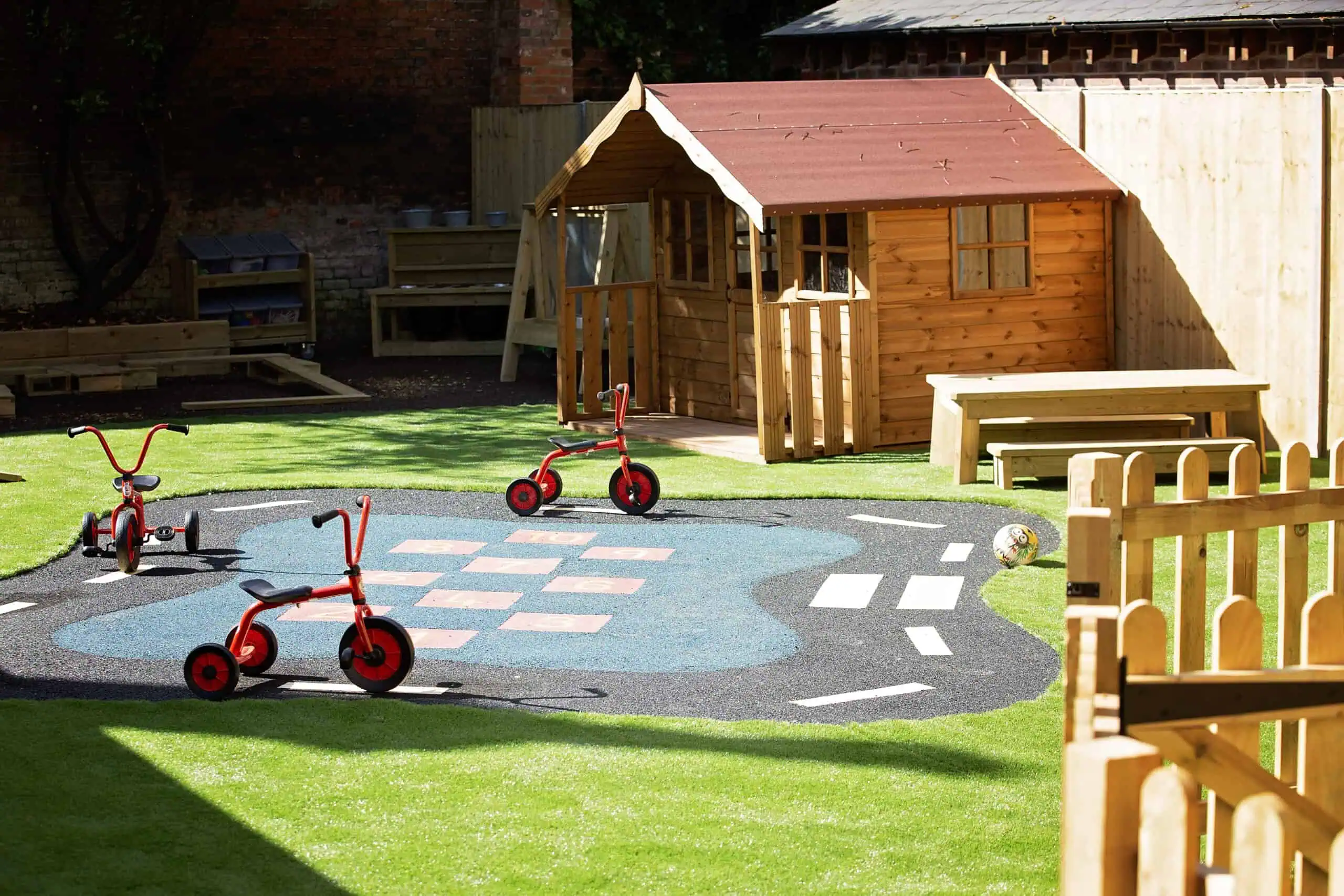 MonkeyPuzzle_Epsom_41 Colorful outdoor play area with tricycles, a hopscotch court, and a wooden playhouse at Monkey Puzzle Day Nurseries.