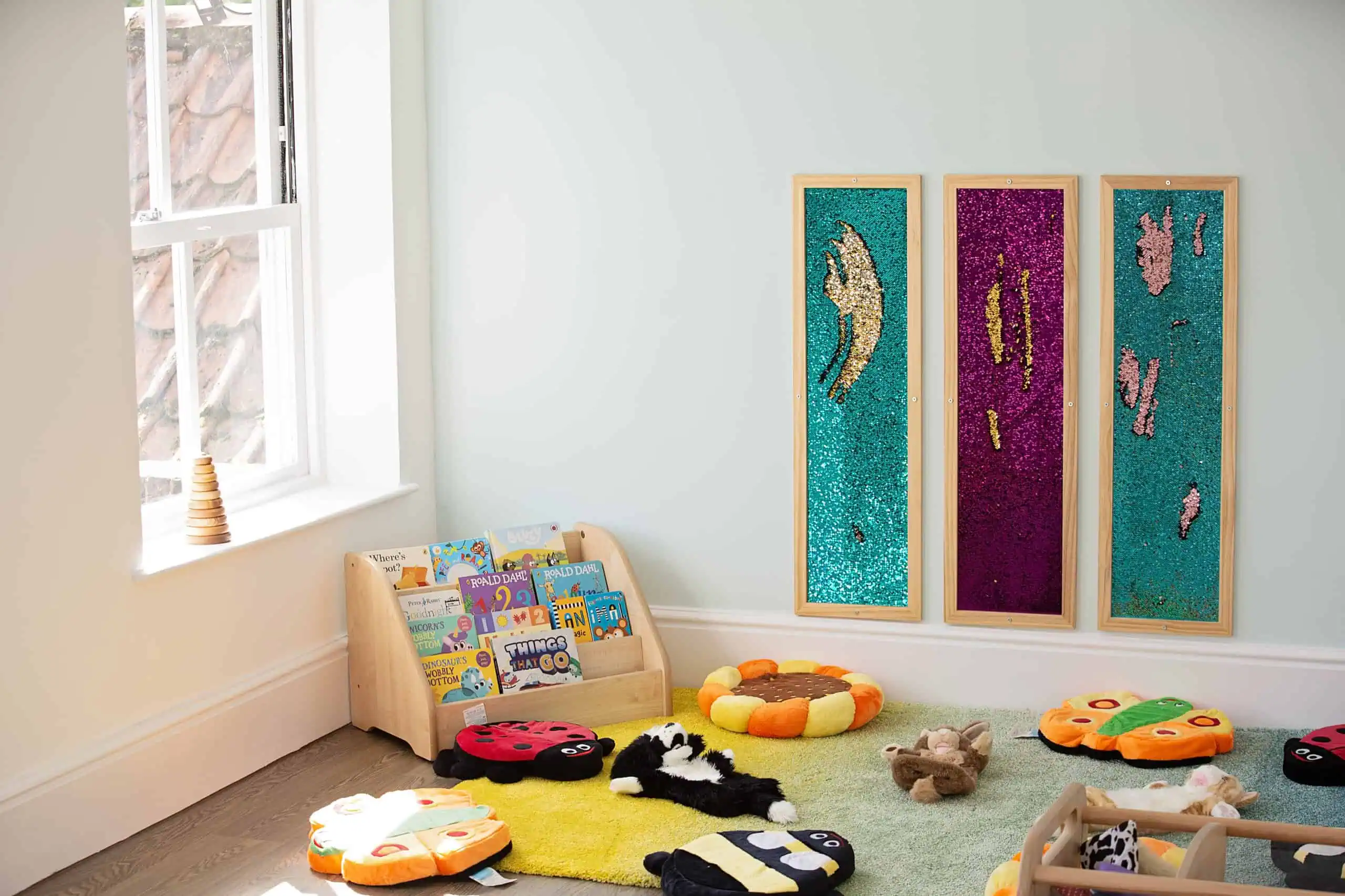 MonkeyPuzzle_Epsom_73 Colorful children's reading nook with low wooden bookcase holding picture books and soft plush toys on a vibrant yellow and green rug, featuring interactive wall displays with sequined art, bright natural lighting from a nearby window, and a welcoming preschool environment at Monkey Puzzle Day Nurseries.