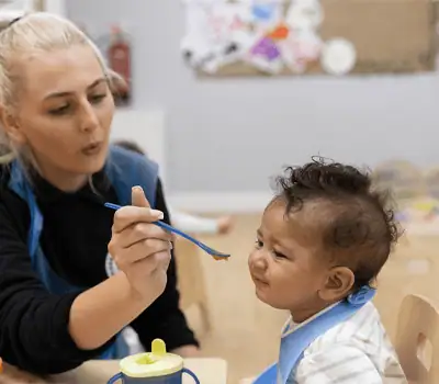 Vibrant scene of a caregiver feeding a young toddler at Monkey Puzzle Day Nurseries, emphasizing nurturing childcare environment and early childhood development in a safe, welcoming nursery setting.