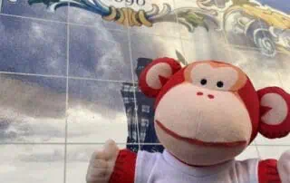 image00003 Monkey Puzzle Day Nurseries mascot plush toy in front of a decorative tile sign reading "Salvor 1890", featuring vibrant colors and playful design. The plush is wearing a "Monkey Puzzle Day Nurseries" T-shirt, creating a welcoming image for early childhood learning environments.