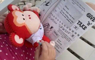 image00011 Cute red and white plush doll held by a child in polka-dot pink clothing, sitting outdoors on a white chair, with a paper activity sheet about giving, taking, swimming, and storytelling, perfect for kids' early learning at Monkey Puzzle Day Nurseries.