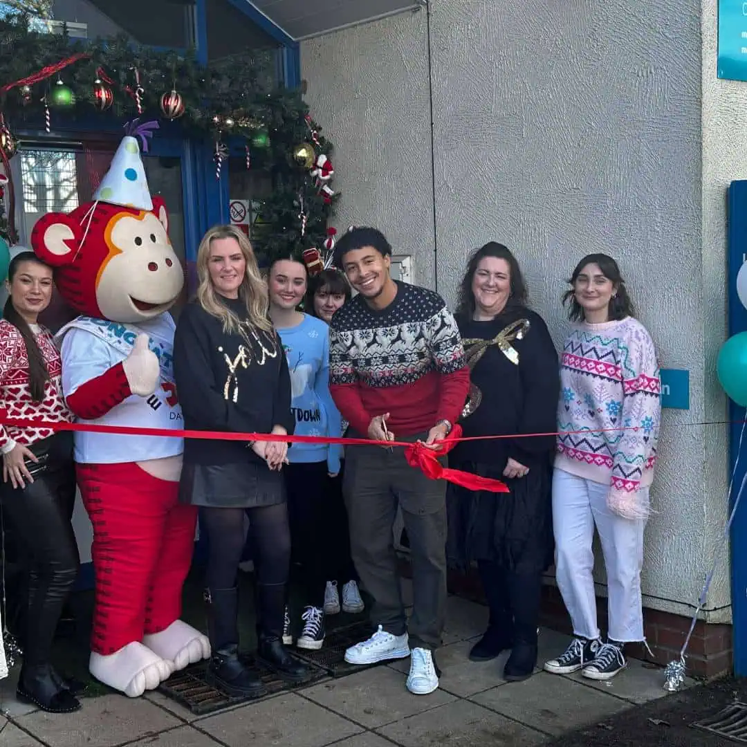 6 Colorful group of teachers and staff celebrating nursery opening with a monkey mascot, festive decorations, and a ribbon-cutting ceremony outside Monkey Puzzle Day Nurseries.