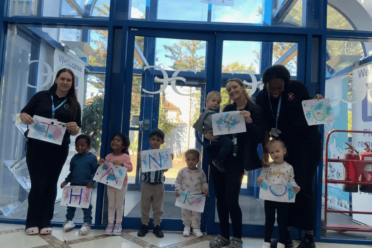 Untitled design (4) Colorful children and teachers holding artwork with the alphabet at Monkey Puzzle Day Nurseries entrance, showcasing a vibrant early childhood education environment focused on learning and development.