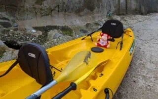 Bright yellow kayak with black seating and weathered paddle on a gravel surface, featuring a red plush monkey toy, outdoor setting near stone wall.