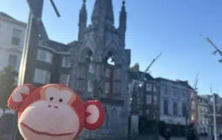 Colorful stuffed monkey toy in front of an iconic cityscape featuring a historic church and a clear blue sky at the Monkey Puzzle Day Nurseries location.