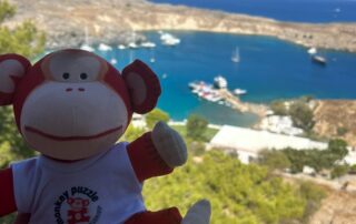 Colorful monkey plush toy with "Monkey Puzzle Day Nurseries" logo, scenic coastal view of boats and hills, perfect for children’s nursery branding and seaside adventure themes.