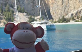 Bright red and white monkey plush toy with "Monkey Puzzle Day Nurseries" logo, sitting by a boat overlooking a scenic lake with sailboats and mountainous backdrop, perfect for early childhood education images.