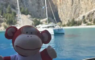Bright red and white monkey plush toy with "Monkey Puzzle Day Nurseries" logo, sitting by a boat overlooking a scenic lake with sailboats and mountainous backdrop, perfect for early childhood education images.